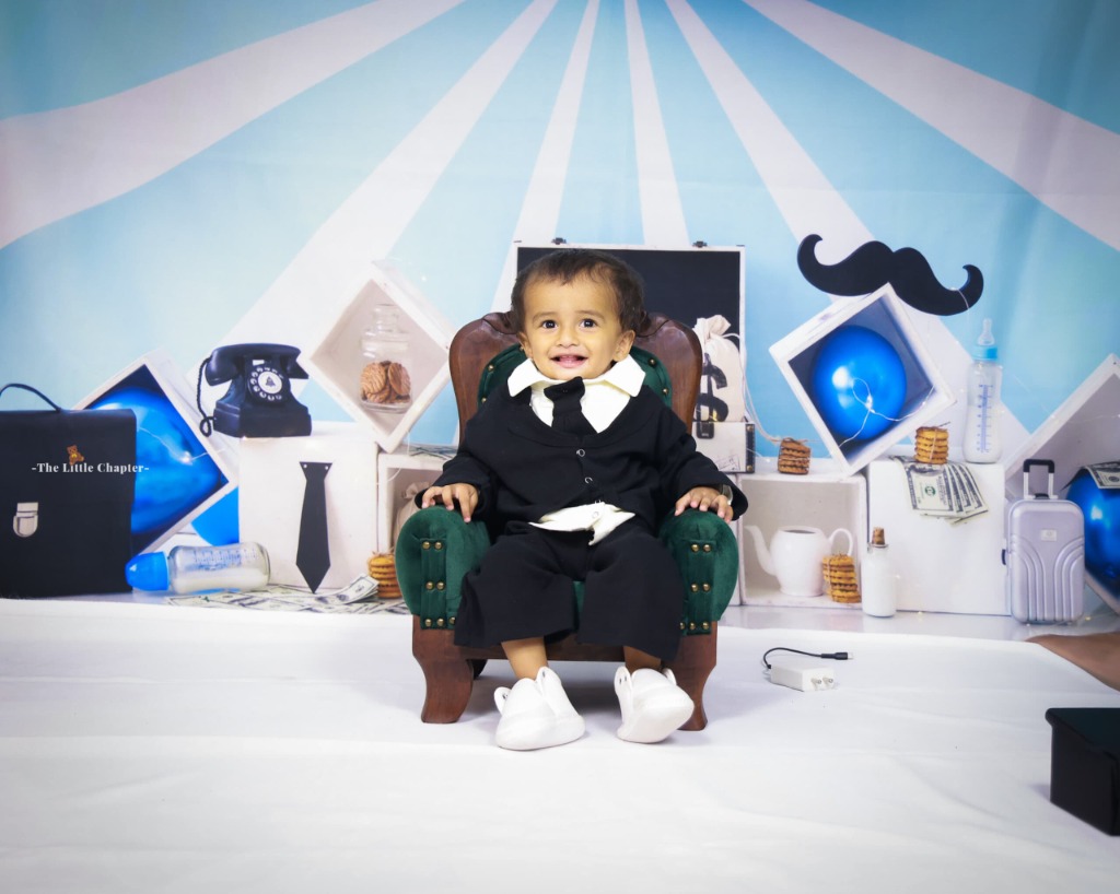 Toddler in black tuxedo suit and tie seated on miniature green armchair with money briefcase