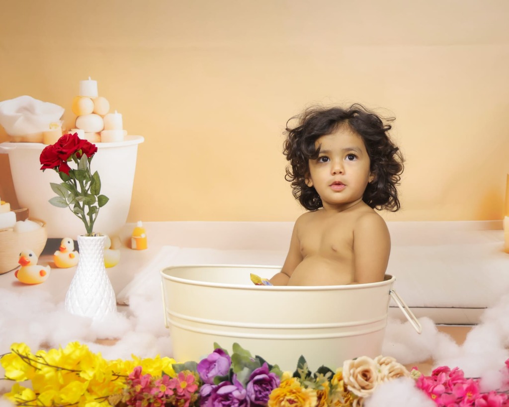 Baby enjoying warm milk bath in vintage ivory tub with colorful flowers and yellow rubber ducks